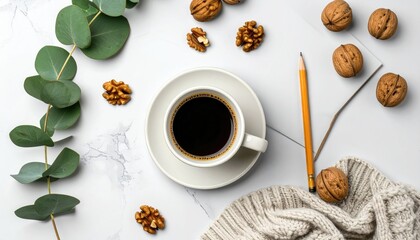 Overhead shot of coffee in a white mug, walnuts, eucalyptus leaves, a pencil, and a cozy sweater on a white marble surface