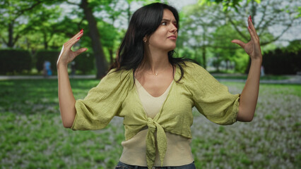 Woman in green top raises both hands at shoulder height in forest under dappled sunlight; confusion...