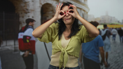 Woman makes hand binoculars gesture at coliseum building under soft daylight; curiosity exploration.