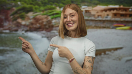 Woman smiling at the seaside beach while pointing outdoors with tattoos, joyfully wearing white and...
