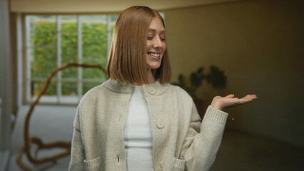 Young blonde woman smiling in a cozy hotel room, wearing a sweater, standing by a large window with...