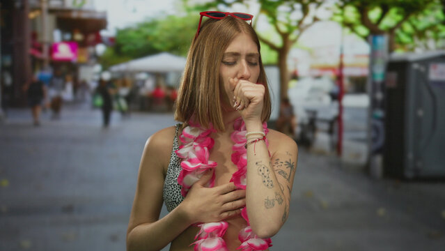 Woman wearing floral lei and sunglasses coughs on city street outdoors, showing expression of discomfort with hand on chest in urban setting.