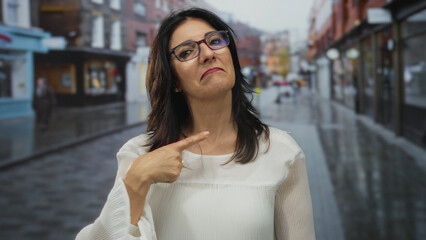 Middle aged hispanic woman points finger to chest on wet urban street with blurred storefronts and reflective pavement; confidence.