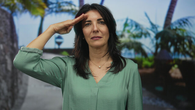 Middle aged hispanic woman raises hand to forehead in forest clearing with palm trunks and lush foliage; respect.