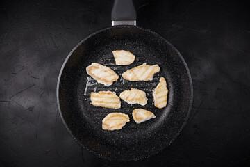 Sizzling pieces of chicken fillet are frying in a non-stick pan on a dark textured surface,...