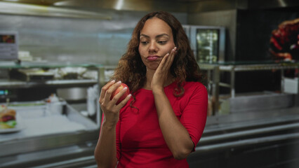 Young african american woman holding an apple, finger on chin, wearing red top by stainless counter...