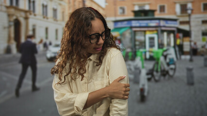 Obraz premium Woman in black framed glasses touching her bare shoulder while wearing beige shirt on crowded city street; quiet discomfort.
