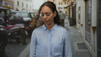 Fototapeta premium Woman with face visible and head bowed while looking down on a narrow urban street with parked scooters and cars; quiet melancholy.
