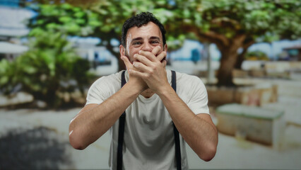 Hispanic man with beard expresses surprise covering mouth with hands outdoors on a street with...