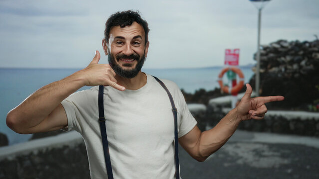 Hispanic man making call gesture on beach promenade with sea backdrop, wearing suspenders and smiling, set against a cloudy sky, evoking casual communication at a seaside location.