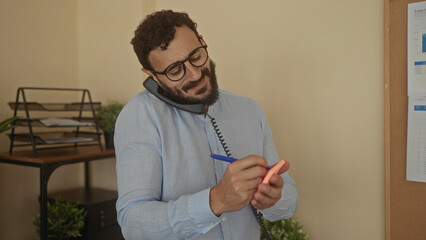 Hispanic man writing notes while multitasking in a modern office setting, holding a phone between his ear and shoulder, creating a busy yet focused work atmosphere.