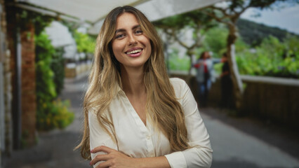 Young blonde woman with long hair crosses arms while smiling under white canopy on garden patio; confidence warmth.