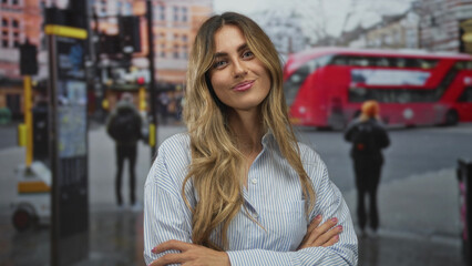 Obraz premium Young blonde woman wearing striped shirt arms crossed smiling at busy city bus stop; confidence friendliness.