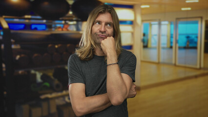 Man points thumb with visible forearm towards dumbbell rack, mirror wall and fitness equipment in studio; hesitation.