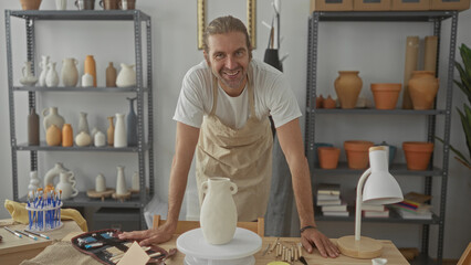 Man wearing apron leaning on wooden table touching a clay vase in a pottery studio lined with...