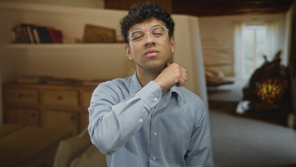 Obraz premium Man wearing glasses holds fist to chest in building interior with warm light and blurred furnishings; determination.