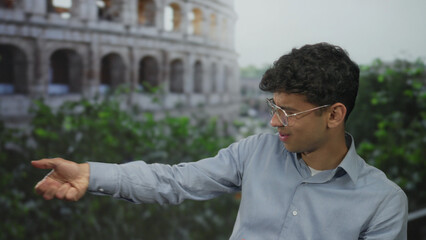 Man gestures with hands toward coliseum building in daylight wearing light blue shirt and glasses;...
