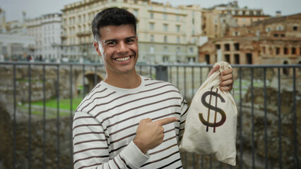 Young man holding money bag in roman ruins backdrop, smiling and pointing, wearing striped shirt,...