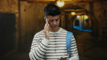 Obraz premium Young man standing on city street wearing striped shirt with blue backpack holding smartphone looking frustrated during night.