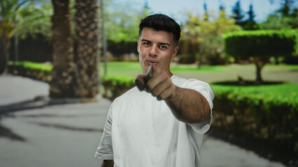 Young man pointing with confidence in an outdoor park with vibrant green trees and sunlight casting shadows, expressing a positive and engaging attitude in a natural setting.