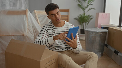 Young man sitting in a new home surrounded by moving boxes while using a tablet in the living room with a relaxed expression on his face.