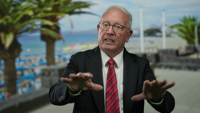 Senior man in suit gesturing on seaside promenade with palm trees and ocean view, conveying a business message outdoors in a calm, professional setting.
