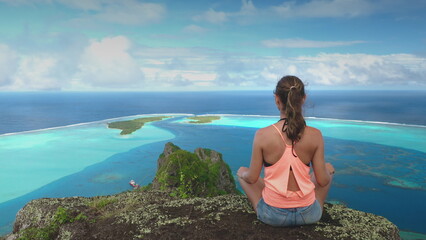 Young woman practices yoga and meditation on a clifftop overlooking the beautiful blue lagoon and motu of maupiti island in french polynesia