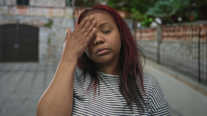 Woman touches forehead wearing wooden watch on cobblestone street outside brick building lined with greenery; stress.