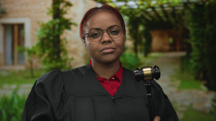 African american woman judge wearing black robe holds gavel outside building with firm posture;...