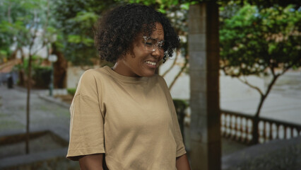Woman grimaces on city street with leafy trees and lush foliage visible while wearing clear...