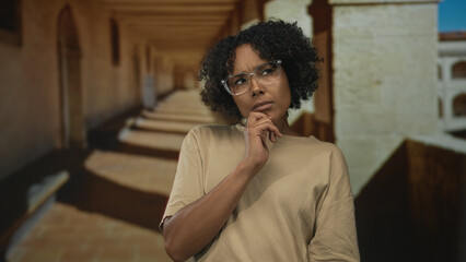 Fototapeta premium Woman in beige shirt and clear glasses rests chin on hand under arched corridor of old stone building; contemplation reflection introspection doubt.