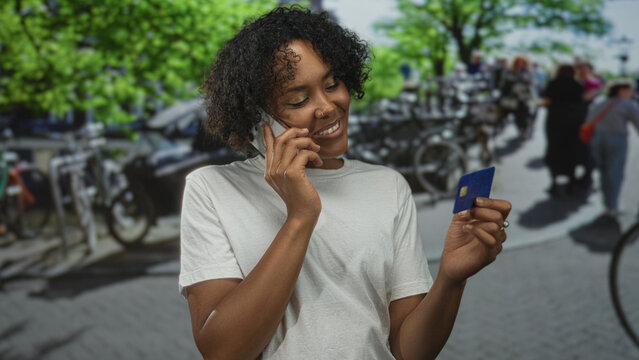 Woman holds smartphone to ear and blue creditcard on sunlit street lined with bicycles and leafy trees; satisfaction.