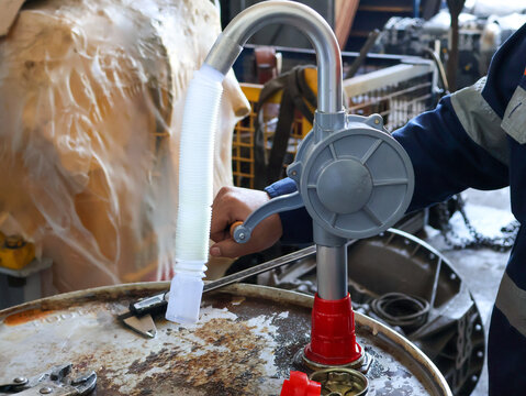 A close-up shot of a worker's hands using a hand pump to transfer fluid from an industrial drum. The image focuses on the tool and the process in an active works