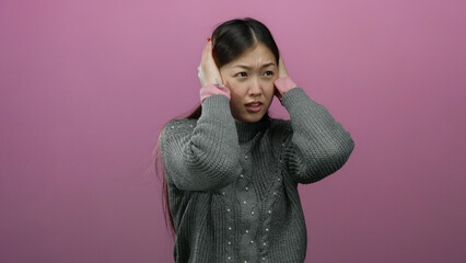Woman covering ears isolated against pink background wearing gray sweater with expressive emotion related to noise or stress showcasing beautiful asian adult female portrait.