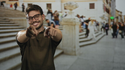 Fototapeta premium Man points finger at camera on crowded street steps near a building facade, smiling and wearing glasses; playful invitation.