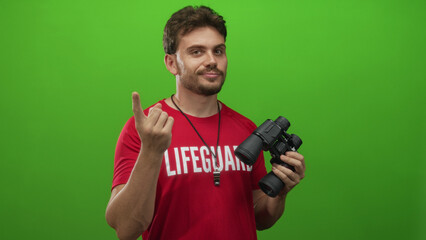 Lifeguard man, young hispanic, pointing finger holding binoculars in green studio; watchful safety...