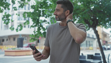Man touching neck and holding smartphone on a tree lined city street with buildings and outdoor...