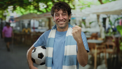 Hispanic man celebrating outdoors on a street, holding a soccer ball and counting on fingers with a big smile, capturing his passion for sports and the vibrant urban atmosphere.