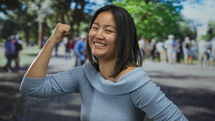 Young chinese woman smiling confidently on a bustling city street, raising her arm in a gesture of...