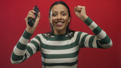 Young hispanic woman with television controller over isolated red background in an expressive gesture conveying excitement and enthusiasm.
