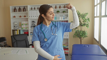 Hispanic female doctor confidently flexes her arms in a hospital room, showcasing strength and...