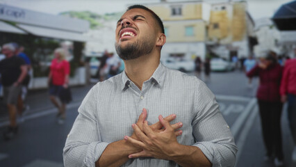 Young hispanic man clutch chest with grimace for heartattack on crowded street in bright daylight;...