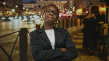 Fototapeta premium Young african american woman wearing glasses with subtle smile and arms crossed on city street at night; rejection.