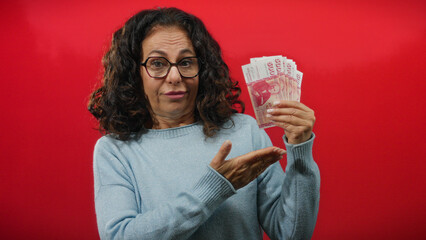 Woman holding icelandic banknotes on isolated red background, expressing curiosity with middle age...