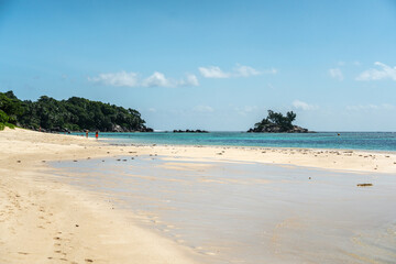 Wide shallow beach in southern Mahe with distant tourists walking and piles of grass