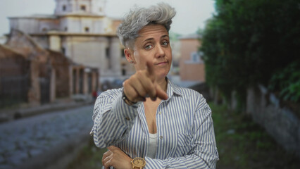 Fototapeta premium Woman with gray hair in striped shirt finger pointing forward on street; doubt uncertainty reflection.