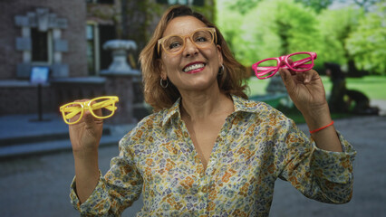 Woman wearing beige eyeglasses holds yellow and pink glasses frames on street showing smiling face;...