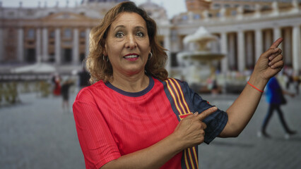 Middle age hispanic woman wearing a red t shirt points finger at fountain in building; joy travel memories.