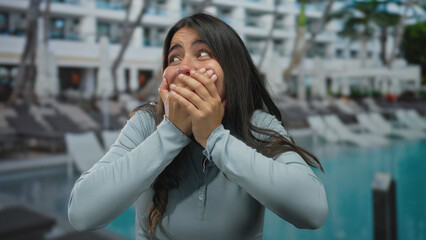 Obraz premium Woman surprised at resort pool with hands covering mouth outdoors showcasing joyful reaction against a hotel backdrop