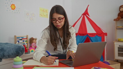 Hispanic woman writing with pen on open notebook beside a laptop on red preschool table in school...